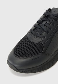 Pantofii sport negri au un upper din plasă și accente din piele netedă, având vârful rotund, șireturile plate și talpa din cauciuc texturat.