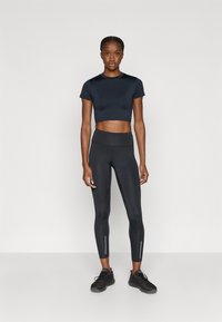 Camiseta corta negra combinada con leggings negros de talle alto que presentan detalles reflectantes. La modelo lleva zapatillas deportivas negras.
