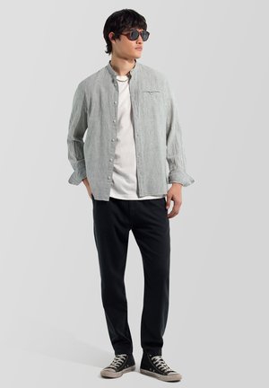 Junger Mann mit runden Sonnenbrillen, weißem T-Shirt, grau gestreiftem Hemd mit Knöpfen, schwarzen Hosen und schwarzen hohen Sneakers, steht mit den Händen in den Taschen.