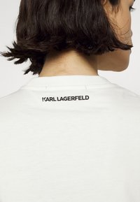 Bílý bavlněný tričko s kulatým výstřihem. Černé logo "KARL LAGERFELD" našité na zadním výstřihu. Hladká textura s uvolněným střihem.