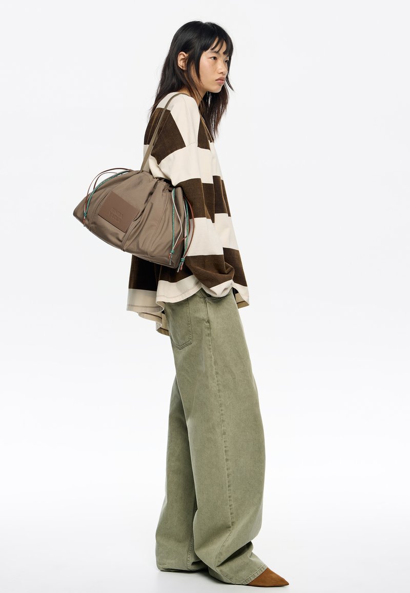  blusa de rayas marrón y crema de gran tamaño, combinada con pantalones de pierna ancha verde claro. Sostiene un bolso marrón con cordones coloridos.