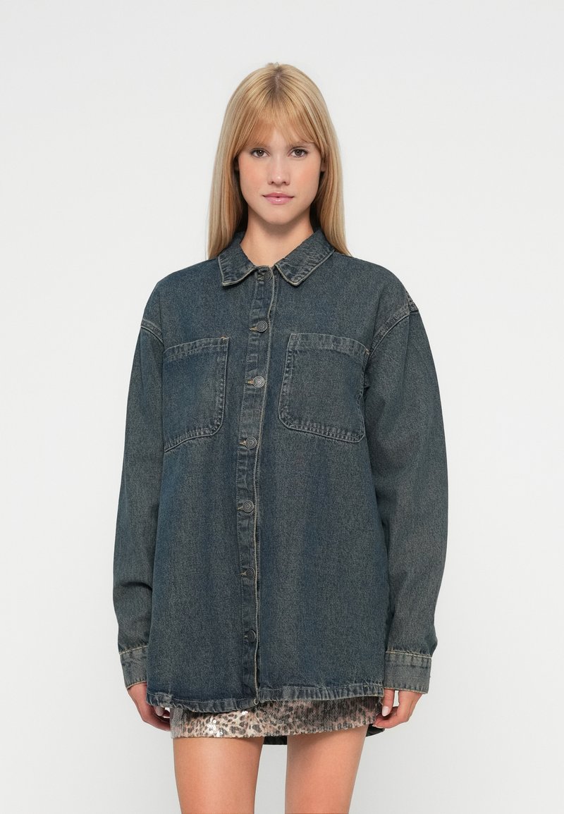 Temna modra denim srajca z dolgimi rokavi, zapenjanjem na gumbe spredaj, dvema žepoma na prsih in sproščenim, ohlapnim krojem. Mehka, teksturirana tkanina.
