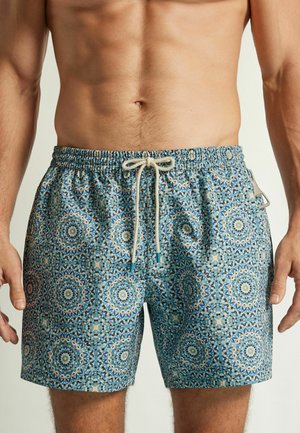 Nahaufnahme des Oberkörpers eines Mannes, der gemusterte blaue Badeshorts mit weißem Kordelzug und geometrischem Mosaikdesign trägt.