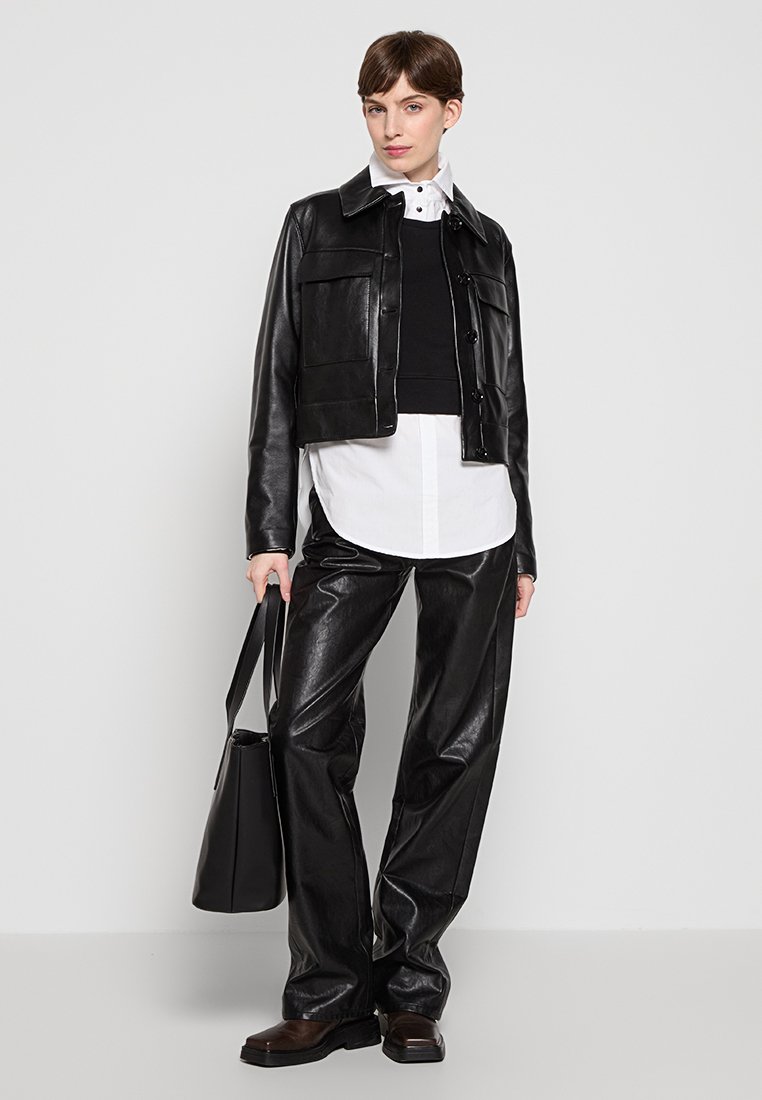 Veste en cuir noir raccourcie avec des boutons, associée à un pull noir, une chemise blanche et un pantalon large en cuir noir. Sac fourre-tout noir.
