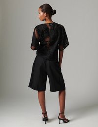 Μαύρο διάφανο τοπ με υφασματικό κυματιστό μοτίβο, λεπτομέρεια με άνοιγμα στην πλάτη· συνδυασμένο με μαύρο, ραμμένο σορτς και πέδιλα με λουράκια.