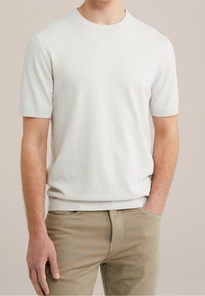 Mann trägt einen weiß gestrickten Kurzarmpullover und beige Hose, steht mit einer Hand in der Tasche vor neutralem Hintergrund.