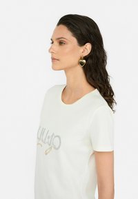 T-shirt blanc à manches courtes avec un texte brillant. Boucles d'oreilles en forme de cœur dorées. Cheveux bruns ondulés. Fond neutre.