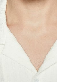 Camicetta bianca con tessuto texturizzato, caratterizzata da un design a V e un solo bottone nel colletto. Pelle visibile sotto il collo.
