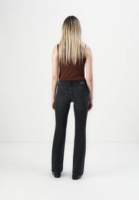 Vrouw met lang blond haar, draagt een bruin mouwloos topje en zwarte jeans, staat met haar gezicht naar een witte muur en haar armen langs haar zij.