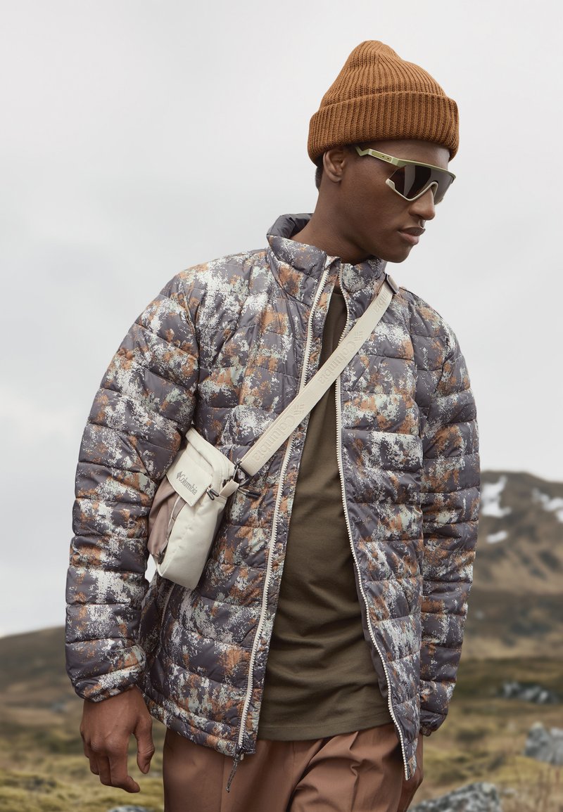 Kurtka pikowana w kolorze szarym z abstrakcyjnym pomarańczowym i białym wzorem; zestawiona z brązową czapką beanie i beżową torebką na ramię.