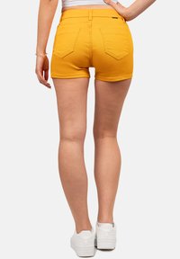 Vrouw in mosterdgele denim shorts, witte sneakers en een witte crop top, staand met één hand op de heup, achteraanzicht.