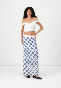 Hvit off-shoulder crop top og et langt skjørt med diamanter i blått og hvitt heklet stoff, med bølgekant. Sorte sko.