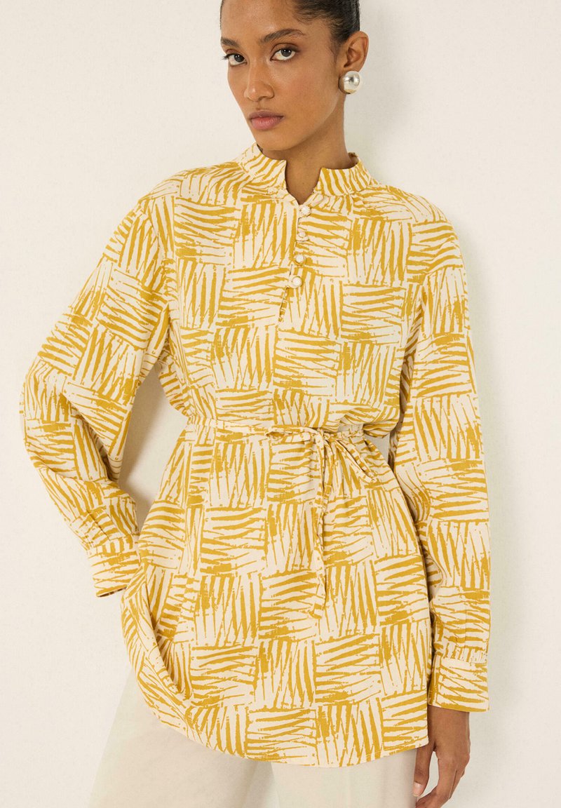 Femme portant un chemisier jaune et blanc à manches longues avec un petit col montant, boutonné sur le devant, et une ceinture à la taille, associé à un pantalon clair.
