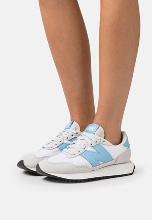 Ben iført hvide og lyseblå New Balance-sneakers med ruskindsdetaljer og sorte gummisåler mod en hvid baggrund.