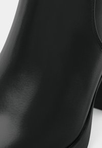 Botte cheville en cuir noir avec une finition lisse et brillante, bout arrondi et talon bloc robuste, présentant des détails de couture subtils.