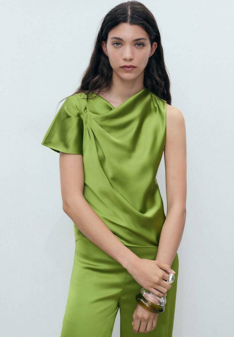 Top en satin vert avec un design à une épaule et des plis, accompagné de pantalons en satin assortis. Le mannequin tient un accessoire en verre transparent.