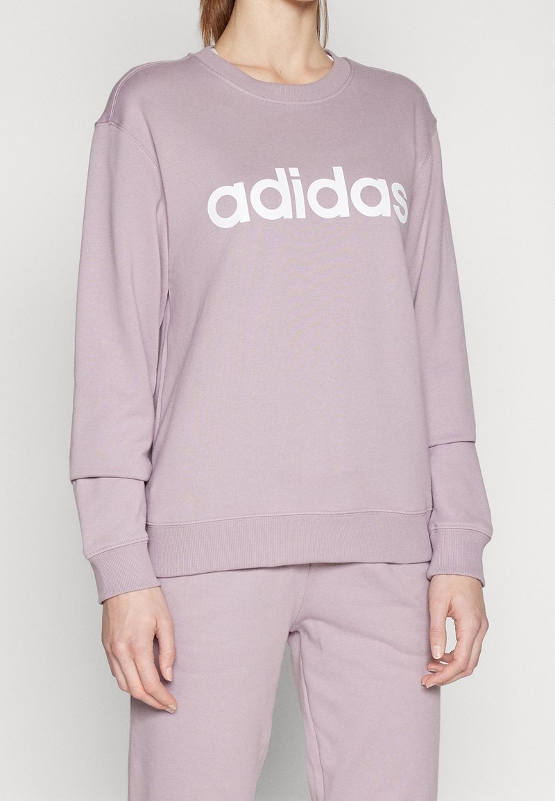 Fialová mikina se kulatým výstřihem a dlouhými rukávy. Na přední straně je výrazné bílé logo "adidas". Měkká, strukturovaná tkanina.