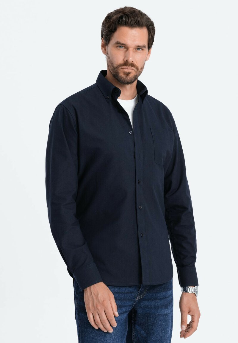 Camisa de botones negra con cuello, un bolsillo en el pecho y mangas largas. Lleva puesta una camiseta blanca debajo, combinada con vaqueros azules.