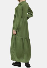 Grünes Maxikleid aus glattem Stoff, mit langen Ärmeln, einem gestuften Design und lockerem Schnitt, kombiniert mit schwarzen Stiefeln.