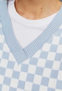 Chaleco de punto a cuadros azules y blancos con cuello en V y ribete, superpuesto a una camiseta blanca. Textura suave, tela blanda.