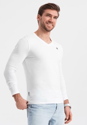 Langärmliges weißes V-Ausschnitt-Hemd aus Baumwollstoff, mit einem kleinen Logo auf der Brust und einer taillierten Passform. Kombiniert mit hellblauen Jeans.