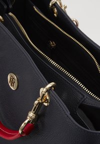 Granatowa skórzana torebka z złotymi akcentami, z zamykanymi na zamek kieszeniami i czerwoną rączką. Teksturowana powierzchnia, przestronne wnętrze.