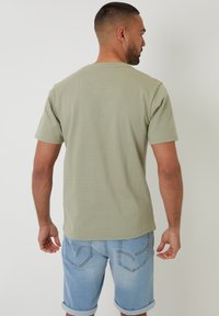 Mężczyzna odwrócony tyłem, ubrany w jasną zieloną koszulę z krótkim rękawem i podwinięte jasnoniebieskie szorty dżinsowe na jednolitym białym tle.