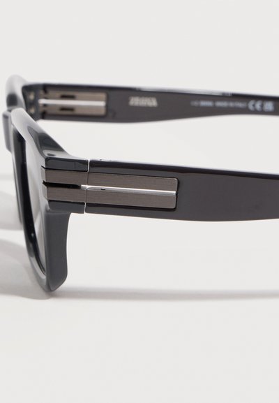 Monture de lunettes rectangulaire noire montrant un gros plan de la branche gauche avec des accents de charnière en métal argenté sur fond blanc.