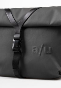 Schwarze wasserdichte Messenger-Tasche mit verstellbarem schwarzen Trageriemen, ausgestattet mit einem Schnallenverschluss und einem geprägten Logo "a/u" auf der Vorderseite.