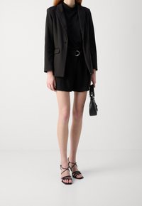 Blazer noir sur un haut noir ajusté, associé à un short noir taille haute. Comprend des sandales noires à lanières et un petit sac à main.