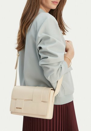 Woman wearing light gray sweatshirt and burgundy pleated skirt, carrying beige crossbody bag with gold logo and bracelet on wrist.