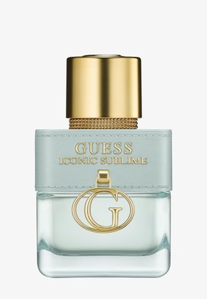 Clear glass perfume bottle with light blue leather band, gold cap, and gold "G" charm labeled Guess Iconic Sublime.