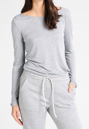 Femme portant un t-shirt gris texturé à manches longues et un pantalon de jogging gris clair à cordon, avec une main dans la poche.