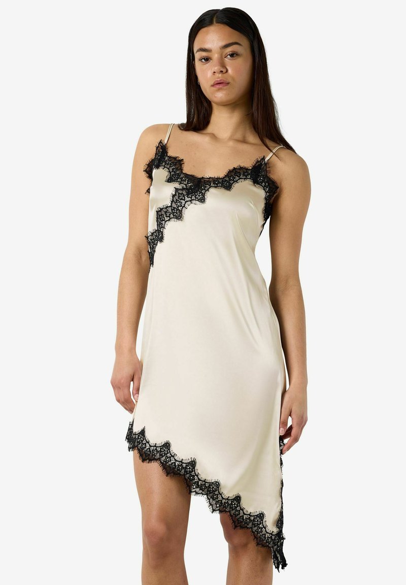 Femme portant une robe nuisette beige asymétrique sans manches avec des bordures en dentelle noire le long de l'encolure, de l'ourlet et du côté, debout devant un fond uni.