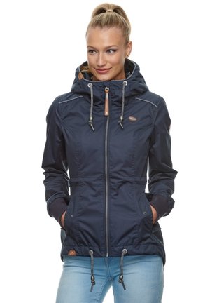 Kobieta z blond kucykiem, ubrana w granatową kurtkę z kapturem i sznurkami, zapinaną na zamek, w jasnoniebieskich dżinsach, z rękami w kieszeniach.