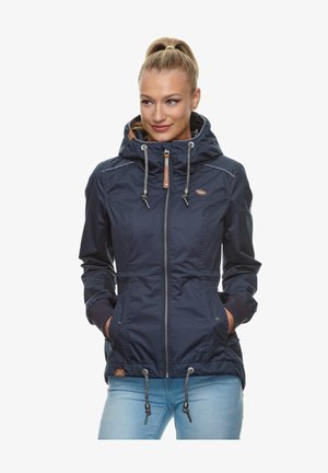 Mujer con coleta rubia que lleva una chaqueta azul marino con capucha y cordones, cremallera frontal y vaqueros azul claro, con las manos en los bolsillos.