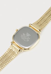 Montre-bracelet en acier inoxydable doré avec un boîtier carré, un bracelet en maille et un dos gris comportant des détails gravés sur la marque et la résistance à l'eau.