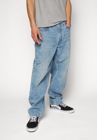 Ljusblå säckiga jeans av denim, med rak skärning och blekt textur. Kombinerade med svarta sneakers med vita skosnören och schackbrädesmönster.