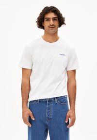 Witte katoenen t-shirt met korte mouwen en een ronde halslijn; voorzien van een blauw tekstlogo op de borst. Gecombineerd met blauwe spijkerbroek.