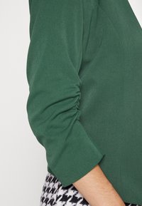 Zelená látková bunda s nařasenými rukávy, která má hladkou texturu. Rukáv je mírně rozšířený a je doplněn černobílou kostkovanou vzorem.