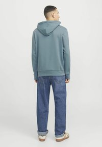 Sudadera con capucha en azul apagado, hecha de una tela suave. Combinada con jeans de mezclilla oscuros de corte holgado y puños, y zapatillas blancas casuales.