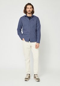 Marine gestreepte overhemdbloes met lange mouwen, gecombineerd met beige broeken en kleurrijke sneakers. Model staat tegen een effen achtergrond.