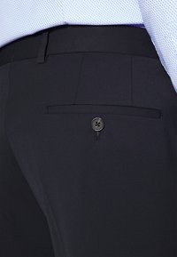 Pantaloni in tessuto nero con tasca posteriore dotata di chiusura a bottone nero, cuciture a vista e una texture liscia.