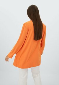 Blusa a maniche lunghe, dal taglio morbido, di un vivace arancione, realizzata in tessuto liscio. Presenta un orlo arrotondato e una leggera caduta, abbinata a pantaloni bianchi.