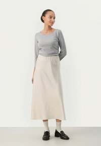 Pitkä beige hame, jossa on joustava vyötärö, yhdistetty tiukkaan harmaaseen pitkähihaiseen paitaan. Mustat loafers-kengät ja vaaleanharmaat sukat täydentävät asustusta.