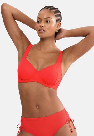 Frau mit geflochtenem Haar, die einen leuchtend roten Bikini mit seitlichen Bändern trägt und mit den Händen hinter dem Kopf vor einfarbigem Hintergrund posiert.