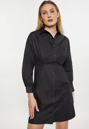 Femme aux cheveux blonds raides portant une robe noire cintrée à boutons avec manches longues sur un fond blanc uni.