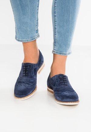 Chaussures brogues en daim bleu marine avec détails perforés, lacets noirs et semelle contrastante couleur beige clair ; associées à un jean bleu clair raccourci.
