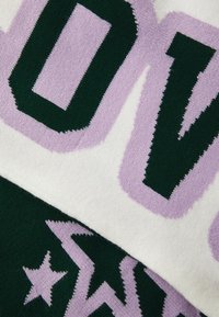 Gestrickte Decke mit großen grünen und lila Buchstaben und Mustern, mit einer strukturierten Optik und kontrastierenden Farben auf einem weichen Hintergrund.