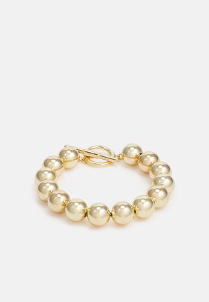 Bransoletka w złotym kolorze z dużymi wypolerowanymi okrągłymi koralikami i zapięciem na toggle, prezentowana na gładkim jasnym tle.